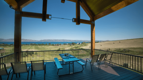 A scenic view from a wooden deck overlooking a lake and rolling hills, with a dining table and chairs in the foreground.